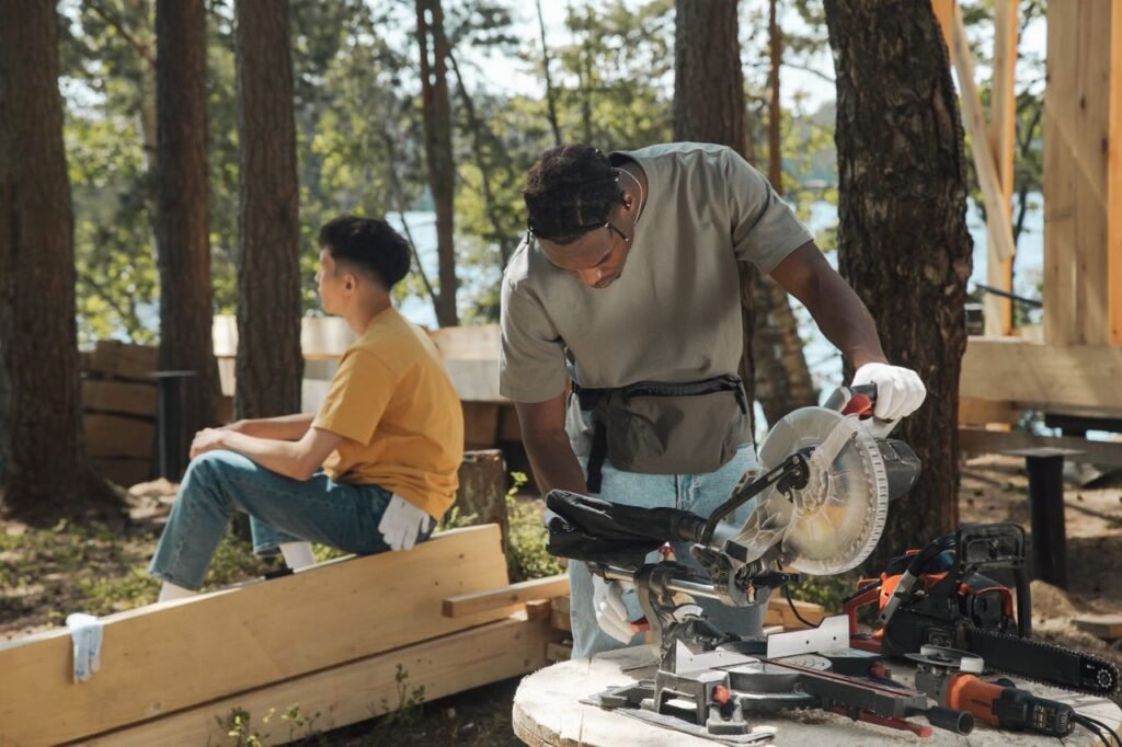 Two men working with carpentry tools on an outdoor construction site in a forest environment.